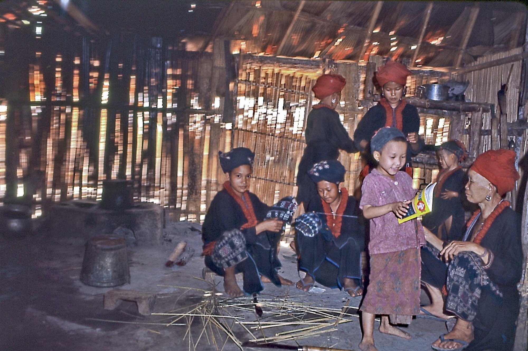 Inside a bamboo house, women in indigo jackets and red ruffs work at embroidery while children stand and read beside an elder.